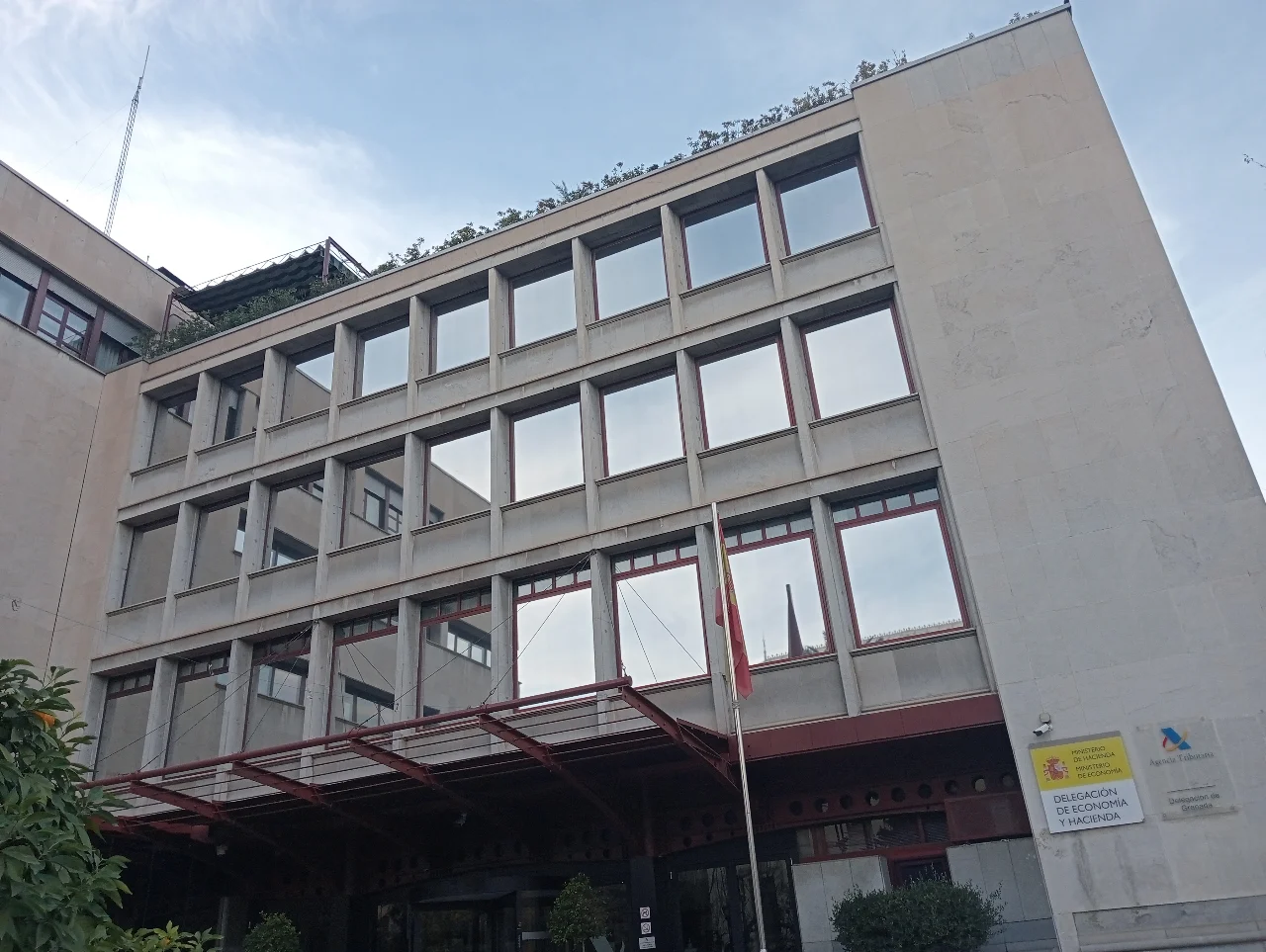 Vista en contrapicado de un edificio institucional con ventanas y banderas; en la fachada se ven los rótulos de la Agencia Tributaria y de la Delegación de Economía y Hacienda de Granada.